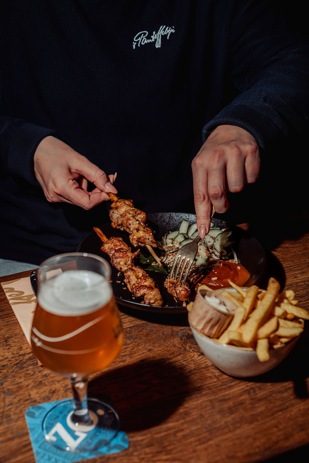 Handen die vlees van een spies halen, naast friet en een glas speciaalbier