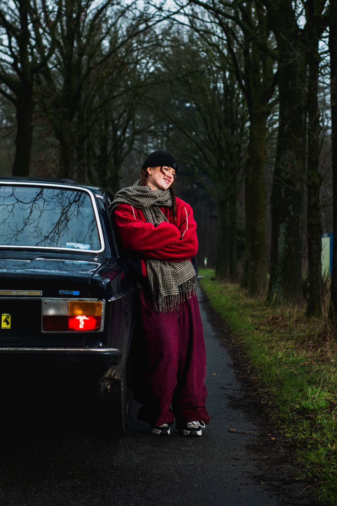 Portret van een man in een rode jas bij een oude auto in een bomenlaan